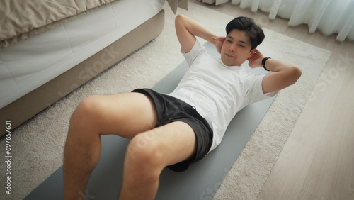 Asian young man doing sit-ups while working out in the home. He is doing workout exercising on sunny morning. Concept of healthy lifestyle and fitness.
