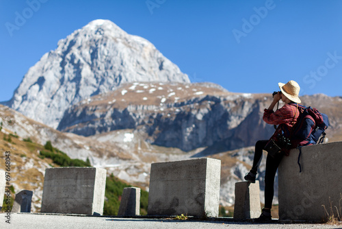 Woman Tourist Sitting on a Concrete Road Block, Taking Photographs of Mount Mangart Beauty in Autumn Colors