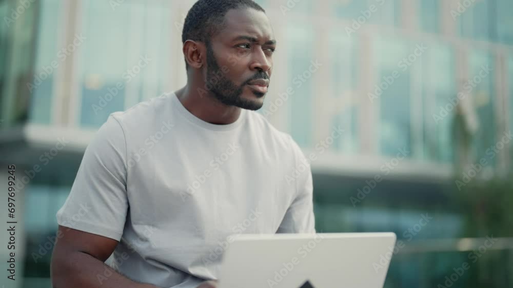 Serious focused man works on laptop sitting on street near modern office building outdoors. African american attentive guy freelancer thinking solving hard work problem online. Freelance work concept. Serious focused man works on laptop sitting on street near modern office building outdoors. African american attentive guy freelancer thinking solving hard work problem online. Freelance work concept.