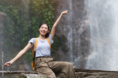 Cheerful young woman with backpack  sitting near waterfall and enjoying the splashing nature power. Traveling and  freedom concept.