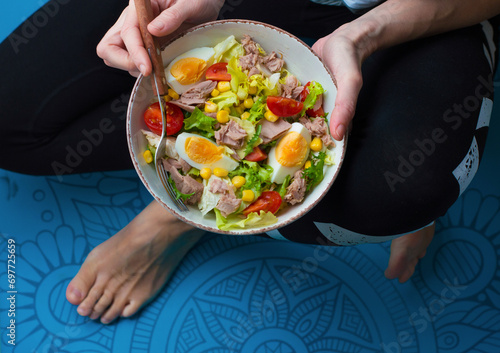 A woman in a sitting position on a blue fitness mat holds in her hands a bowl of salad with tuna and vegetables. Healthy nutrition concept. Horizontal.