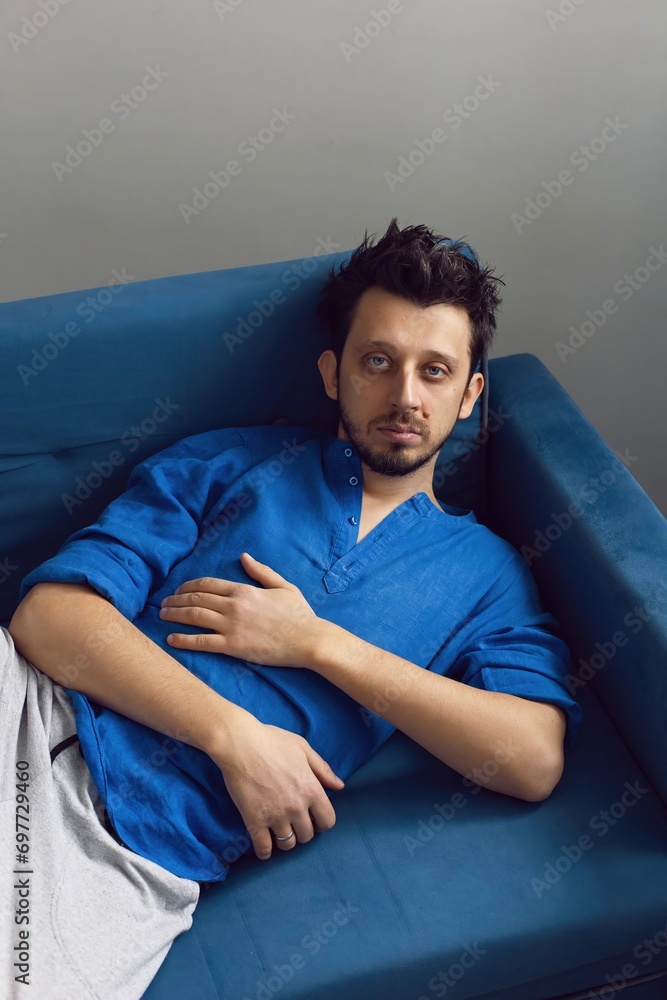 Stylish portrait of a hipster man with a beard and blue eyes in a blue shirt sitting on a sofa in a studio against a white wall. guy 35 years old light from the window Stylish portrait of a hipster man with a beard and blue eyes in a blue shirt sitting on a sofa in a studio against a white wall. guy 35 years old light from the window