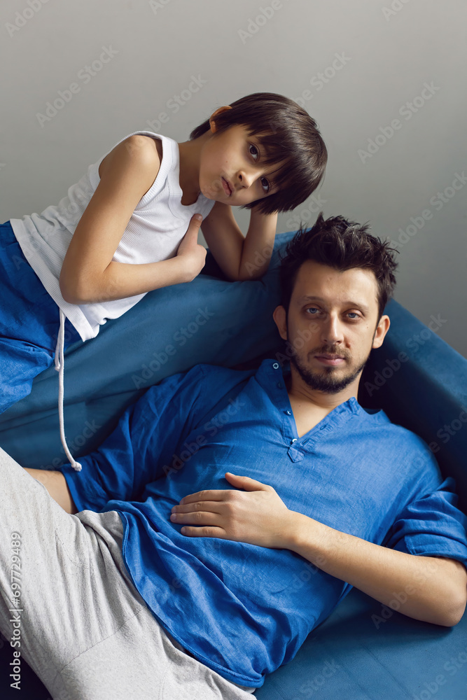 Obraz premium Portrait of a family father and son sitting on a blue sofa in the studio opposite the window. A hipster man with a beard and blue eyes in a shirt hugs his child.