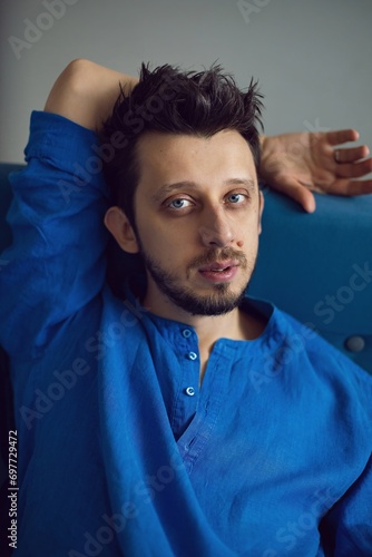 Stylish portrait of a hipster man with a beard and blue eyes in a blue shirt sitting on a sofa in a studio against a white wall. guy 35 years old light from the window