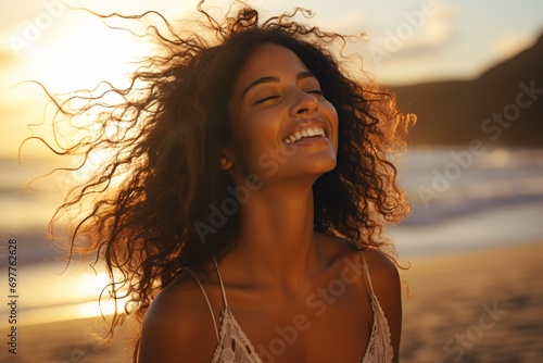 A lovely woman of mixed heritage smiling while relaxing on a beach at sunset with her eyes closed and hair blowing in the wind.