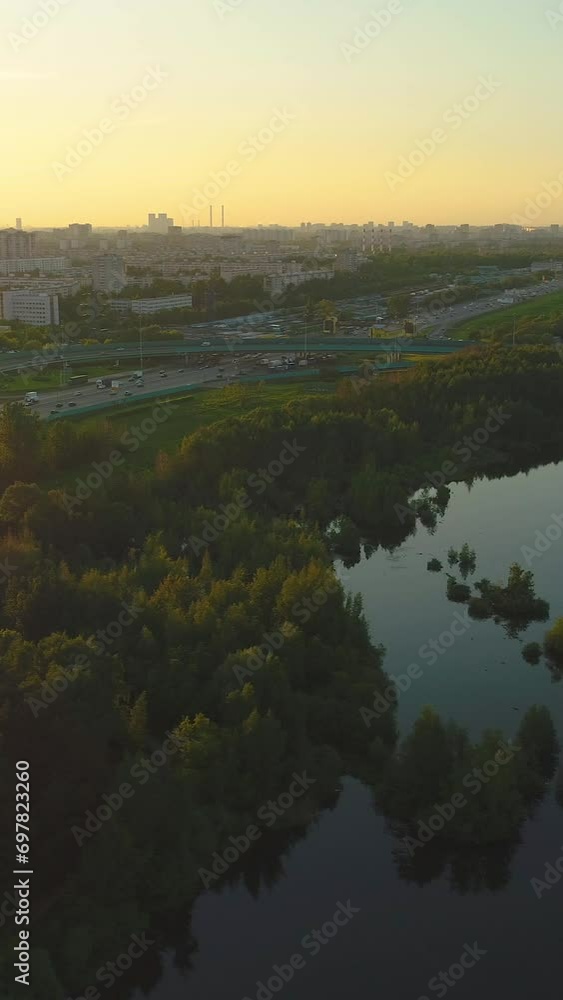 Flying around the lake, trees and highway with cars traffic in the city at sunset. Aerial view. Vertical Video Flying around the lake, trees and highway with cars traffic in the city at sunset. Aerial view. Vertical Video