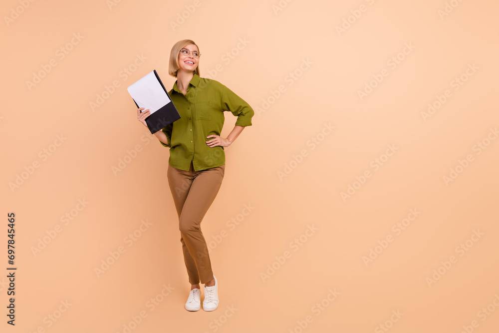 Full size photo of lovely woman dressed green shirt in glasses hold clipboard look at logo empty space isolated on beige color background Full size photo of lovely woman dressed green shirt in glasses hold clipboard look at logo empty space isolated on beige color background