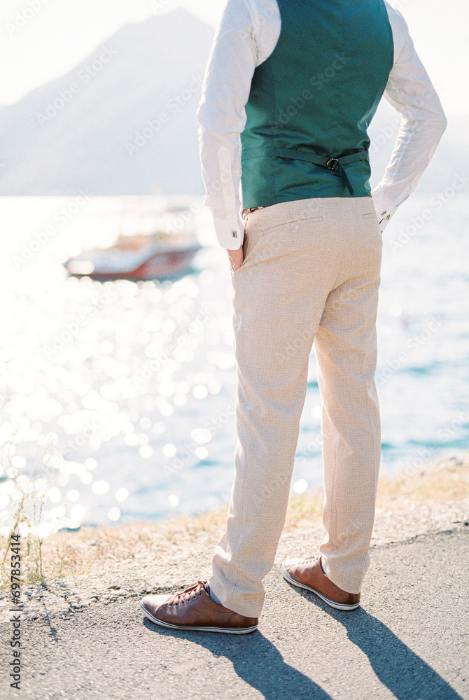 Groom stands on the embankment with his hands in his trouser pockets. Back view. Cropped. Faceless
