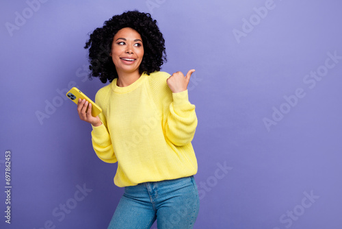 Photo portrait of pretty young girl point look empty space hold device wear trendy yellow outfit isolated on purple color background