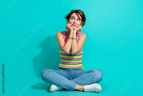Full length photo of gorgeous creative person sit floor arms touch cheekbones look empty space isolated on teal color background