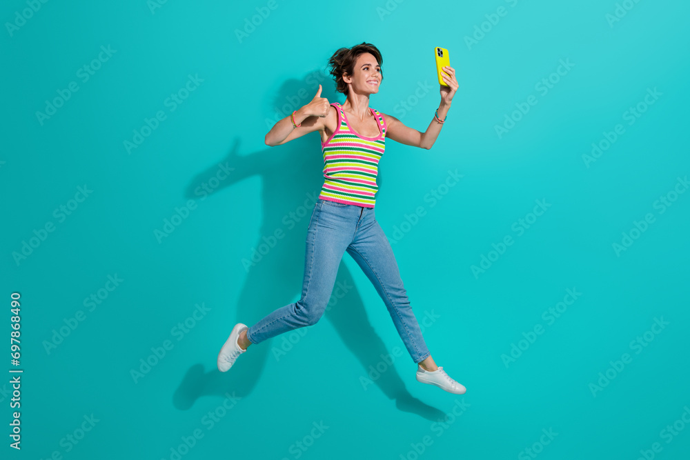 Full size photo of charming sportive girl jump hold smart phone take selfie show thumb up isolated on teal color background