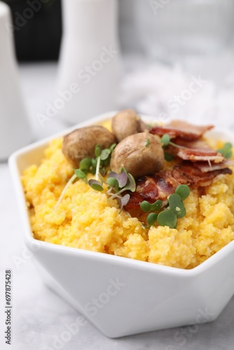 Cooked cornmeal with bacon, mushrooms and microgreens in bowl on white marble table, closeup