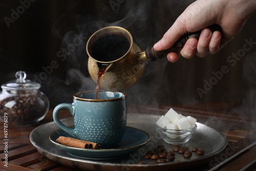 Turkish coffee. Woman pouring brewed beverage from cezve into cup at wooden table, closeup photo