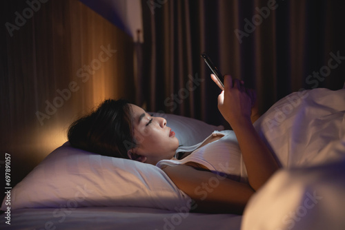 Asian woman using mobile phone smartphone laying on the bed in the bedroom. Sleepy exhausted, can not sleep. Insomnia, addiction concept. Women scrolling social networks on mobile dark bedroom. photo