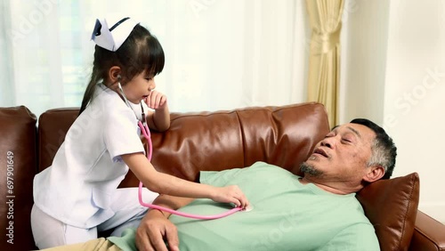 Little nurse plays nurse doctor with grandpa who teases sick lying on the sofa using medical stethoscope to listen to the pulse playing together and having fun.