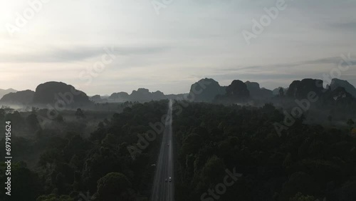 A video from a drone slowly flying from a long straight road with cars moving back and forth in the morning, gradually ascending until it reveals a distant view of forests, mountains, and mist in the 