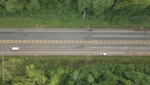The video shows a drone slowly flying from right to left, capturing a wide road with cars driving smoothly while surrounded by lush forests.