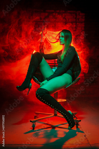 Beautiful girl in bodysuit sits on the iron armchair on the red light smoke background. Beauty and fashion concept.