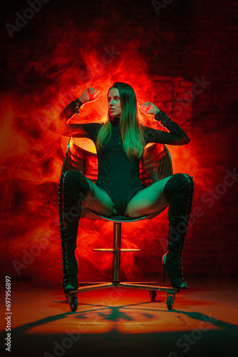 Beautiful girl in bodysuit sits on the iron armchair on the red light smoke background. Beauty and fashion concept.