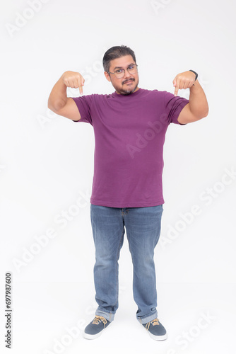 A middle aged man pointing with his fingers to something below or showing his shoes. Wearing a purple waffle shirt and jeans. Full body photo, isolated on a white background.