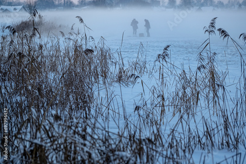 ice and snow covered river with dry reeds and two silhouettes of fishermen in fog. Misty cold winter morning in Latvia