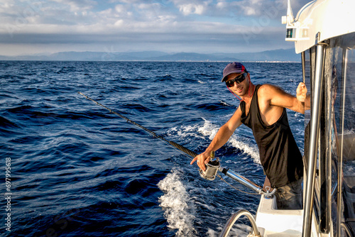 Tough man wearing baseball cap on board ship while fishing against blue sea background.