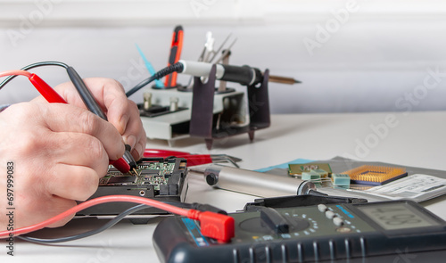 Close-up of a technician's hands in a workshop. The repairer is using a multimeter to measure the parameters of an electronic device on a table.