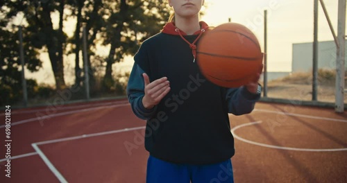 A close-up portrait of a blonde girl in a sports uniform who throws an orange ball from hand to hand on a red basketball summer street court in the morning photo