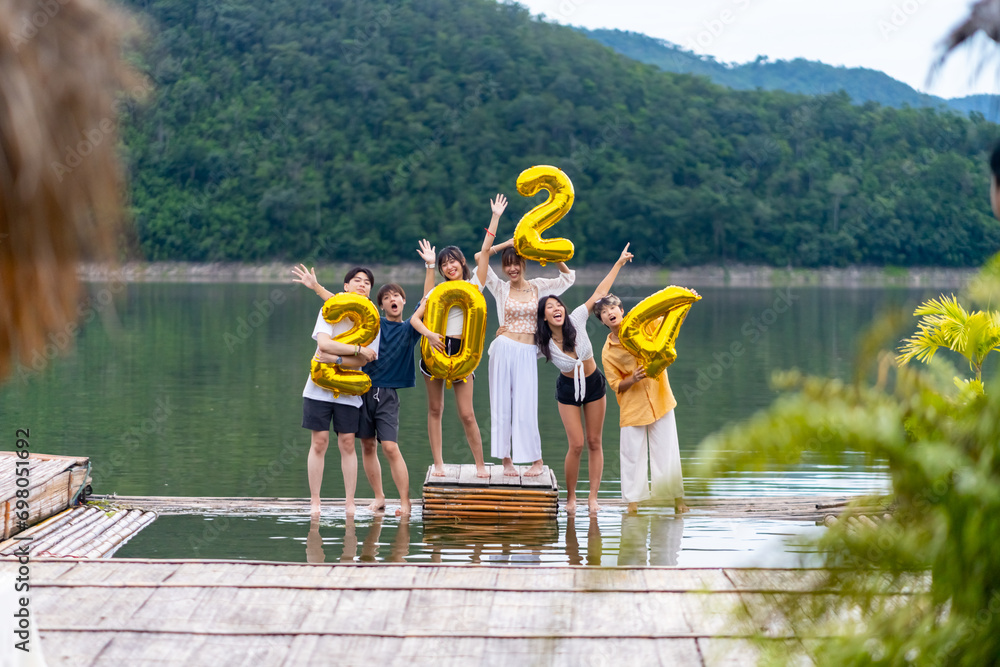 Group of Young Asian man and woman friends celebration happy new year party holding golden number 2024 balloon. People enjoy and fun travel nature mountain together at lake house on holiday vacation. - obrazy, fototapety, plakaty 