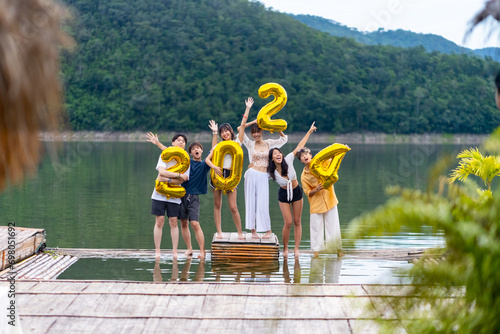 Group of Young Asian man and woman friends celebration happy new year party holding golden number 2024 balloon. People enjoy and fun travel nature mountain together at lake house on holiday vacation.