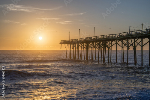Serene dawn at Carolina Beach with a rustic pier stretching into the calm ocean against a pastel sunrise sky. © Red Lemon