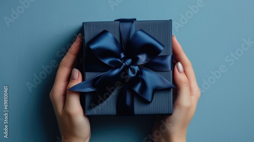 Top view to young woman hands holding navy blue present gift giftbox with satin ribbon bow on light blue background. Christmas, birthday, anniversary, shopping, blue friday concept