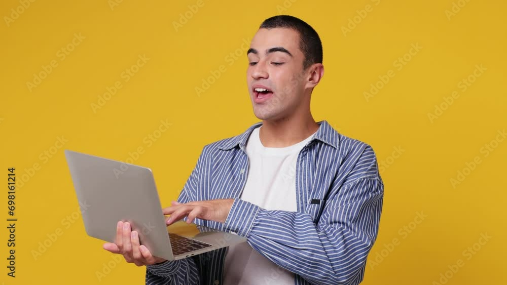 Smart fun IT user young middle eastern man he wear blue shirt white t-shirt hold in hand use work typing on laptop pc computer isolated on plain yellow background studio portrait. Lifestyle concept Smart fun IT user young middle eastern man he wear blue shirt white t-shirt hold in hand use work typing on laptop pc computer isolated on plain yellow background studio portrait. Lifestyle concept