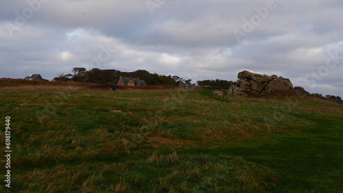 Photo de gros rochers aux bords des côtés de la Bretagne, sous une mer agitée, avec de bruyère ou végétation vert et orange, maritime, ciel gris, nuageux et venteux, environnement celtique, maison 
