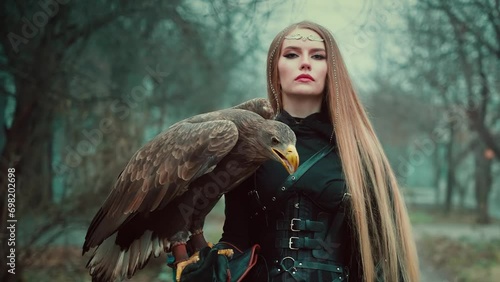 art portrait fantasy woman warrior holding white-tailed eagle wild bird on hand. Princess girl long blond hair. Lady queen assassin hunter black dress armor walking along street dark trees fog. 4k
