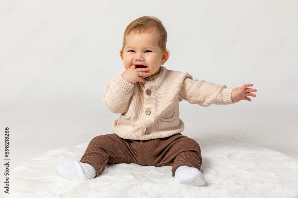 Happy smiling adorable newborn baby girl in beige and brown suit sitting on light white home carpet on white background. 6 to 7 months old development. advertising of toys and goods for children Happy smiling adorable newborn baby girl in beige and brown suit sitting on light white home carpet on white background. 6 to 7 months old development. advertising of toys and goods for children