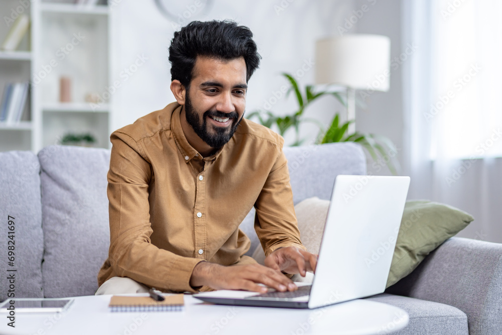 Indian smiling young man sitting on sofa working and studying remotely at home using laptop. Indian smiling young man sitting on sofa working and studying remotely at home using laptop.