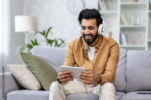 Canvas Print Indian young man sitting on the couch at home wearing headphones and using a tablet with a smile