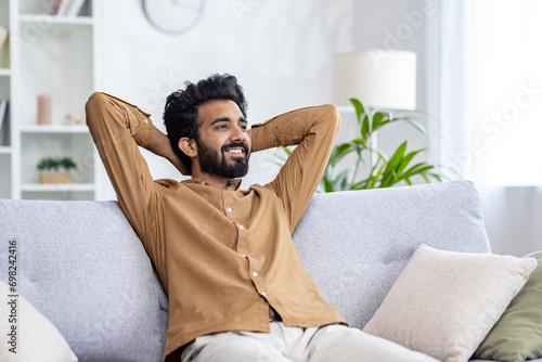 Happy Indian young man sitting relaxed on sofa at home with hands behind head and resting, smiling contentedly. photo