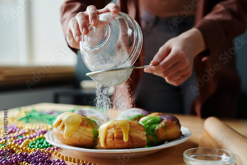 Hands of young unrecognizable woman sieving sugar powder on top of appetizing homemade donuts on plate while preparing for Mardi Gras