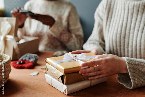 Hands of girl wearing sweater on stack of packed and wrapped Christmas or New Year gifts with tied silk white ribbon on top