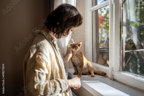 Fotografie Tender loving teenager caressing, stroking adorable breed cat Devon Rex sitting on windowsill, warming in sunlight