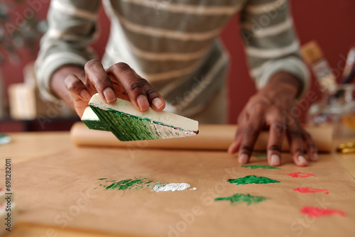 Woman holding handmade firtree workpiece over unrolled craft paper and making prints of Christmas symbols while preparing for holiday photo