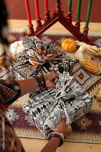 Hands of young unrecognizable woman tying knot on top of packed gift wrapped into headscarf against candlestick and ripe vagetables photo