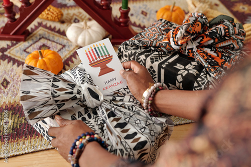 Hand of young African American woman holding creative Kwanzaa postcard with candleholder over packed and wrapped present photo