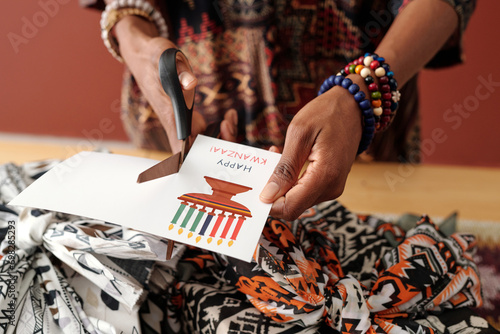 Hands of young unrecognizable African American woman with scissors cutting part of handmade Kwanzaa postcard while preparing for holiday photo