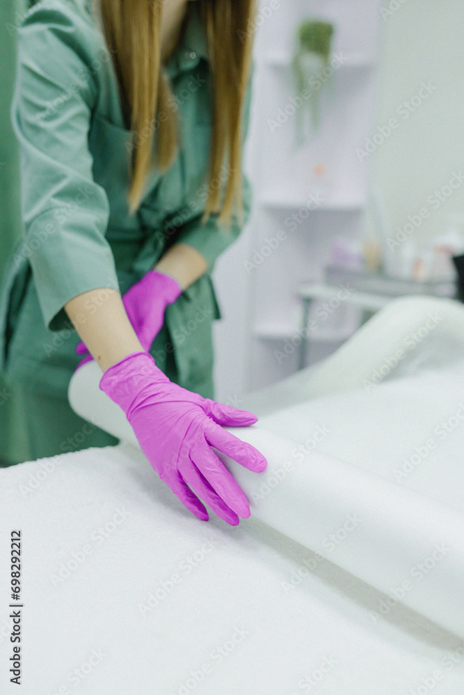 An hair removal master spreads a protective film on the couch for a girl patient before starting a procedure in a beauty salon, a cosmetologist in a cosmetology office loves sterility and cleanliness
