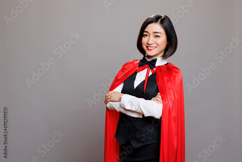 Confident asian waitress wearing hero red cloak, standing with folded hands and showing power portrait. Smiling woman receptionist in superwoman cape looking at camera in studio