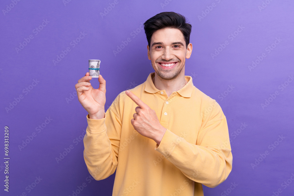 Photo of positive good mood man dressed yellow shirt pointing finger banknotes roll empty space isolated purple color background Photo of positive good mood man dressed yellow shirt pointing finger banknotes roll empty space isolated purple color background