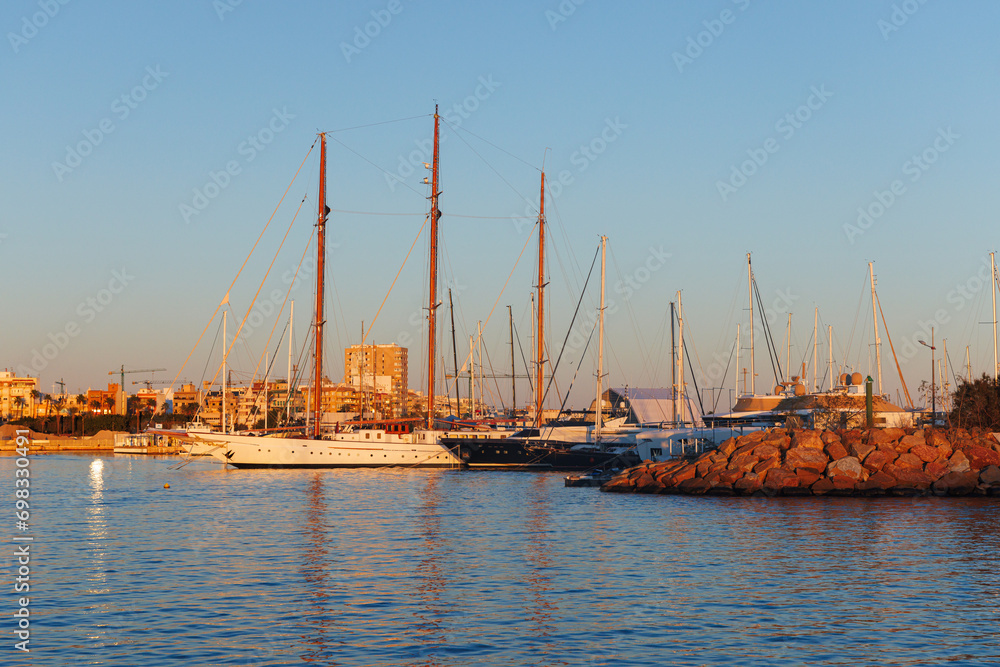 A sailing yacht with a wooden mast stands in the marina at sunset.